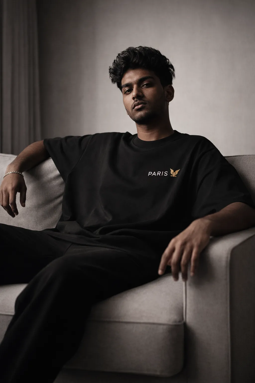 Young Indian male model sitting on sofa wearing Paris Eagle oversized streetwear T-shirt with logo